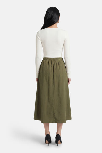 Jupe kaki mi-longue de Le Temps des Cerises, avec taille élastique et coupe évasée, portée avec un haut blanc à manches longues.
