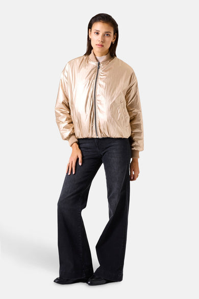 Veste courte beige métallisée de GARCIA, avec fermeture éclair et bords-côtes beiges, portée avec un jean noir évasé.
