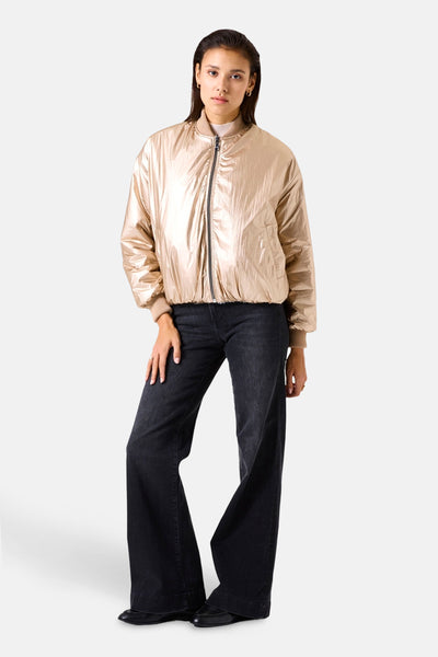 Veste courte beige métallisée de GARCIA, avec fermeture éclair et bords-côtes beiges, portée avec un jean noir évasé.
