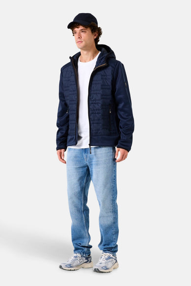 Veste de pluie bleue de GARCIA, avec capuche et fermeture éclair, portée avec un t-shirt blanc, un jean bleu et des baskets.