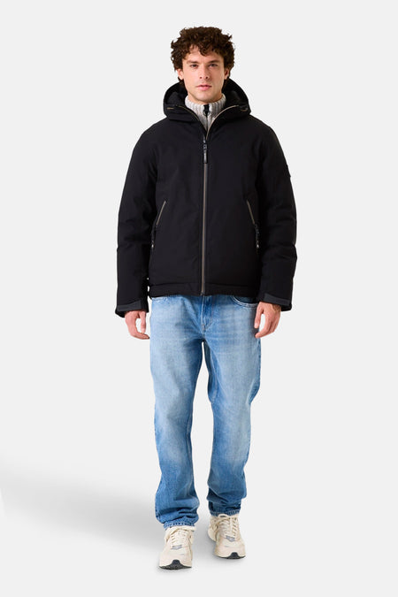 Zwarte regenjas van GARCIA, gedragen met een grijze coltrui, een blauwe spijkerbroek en witte sneakers.