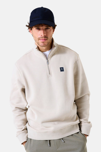 Beige sweater met opstaande kraag van GARCIA, met ritssluiting tot halverwege en vierkant logo, gedragen met een marineblauwe pet en een joggingbroek.