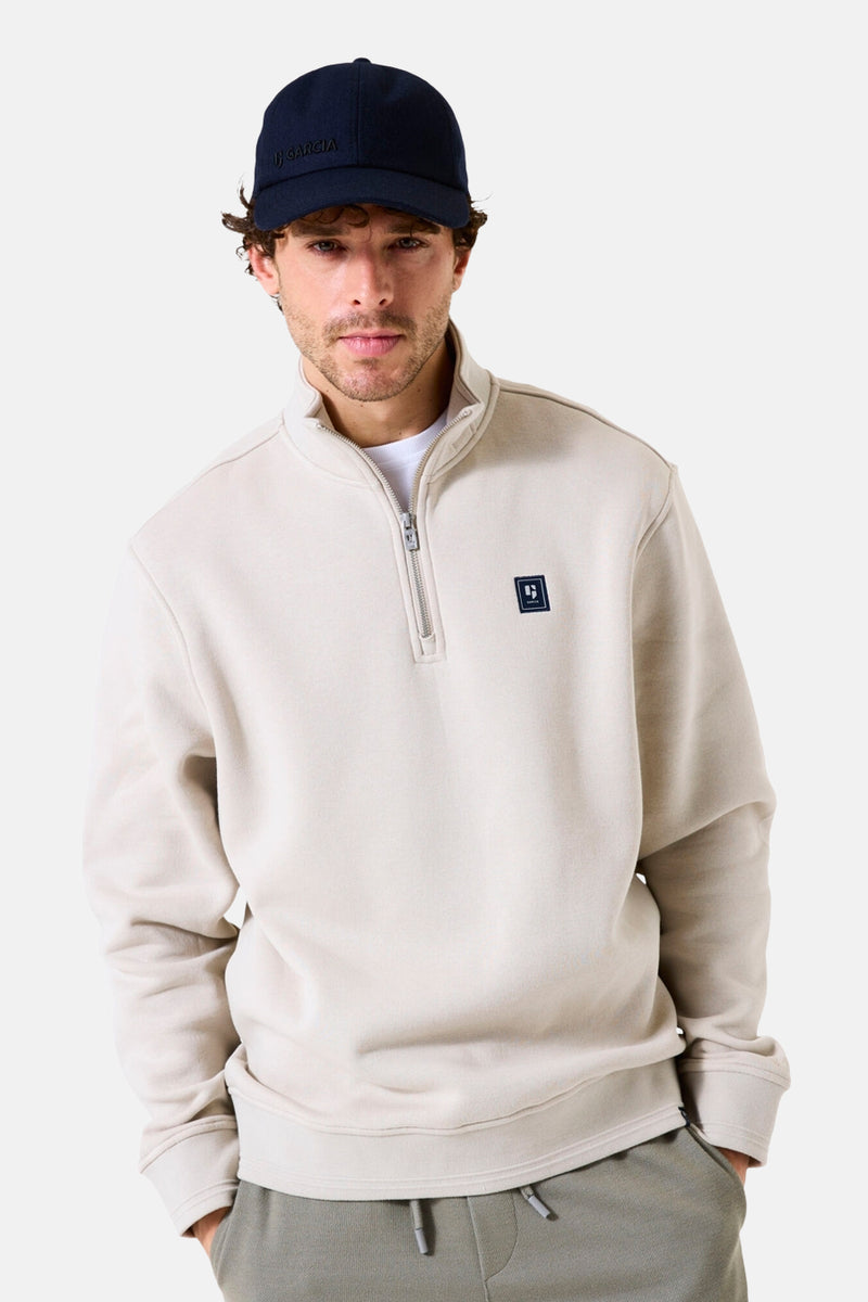 Beige sweater met opstaande kraag van GARCIA, met ritssluiting tot halverwege en vierkant logo, gedragen met een marineblauwe pet en een joggingbroek.