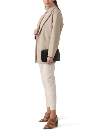 Zwarte Guess schoudertas, gedragen met een beige jas, een trui en gebroken witte broek, en bruine hoge hakken.