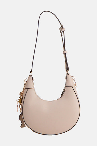 Sac à bandoulière beige de Guess®, avec une bandoulière noire réglable et des breloques dorées décoratives.
