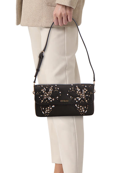 Zwarte Guess schoudertas versierd met glinsterende bloemenpatronen, gedragen met een beige jas en een witte broek.