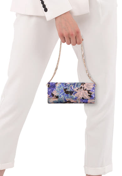 Multikleuren schoudertas van Guess, versierd met pailletten met paarse, blauwe en roze abstracte patronen, vastgehouden door een hand in een witte outfit.
