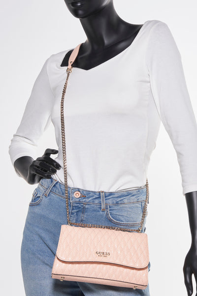Sac à bandoulière rose poudré de Guess, avec bandoulière chaîne, sur mannequin portant un jean et un T-shirt blanc.
