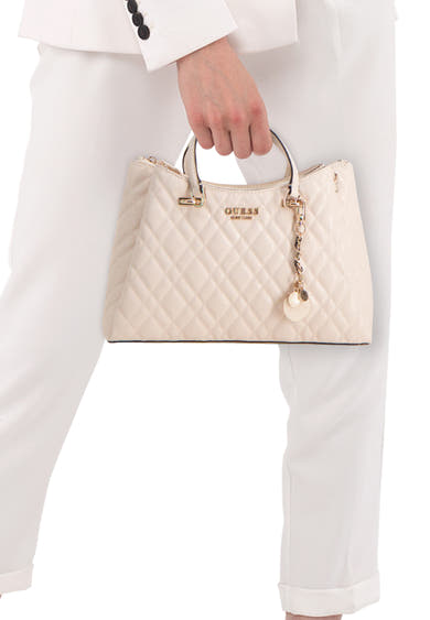 Beige gewatteerde handtas van Guess, met decoratieve bedel en gedragen door een persoon in een wit broekpak.
