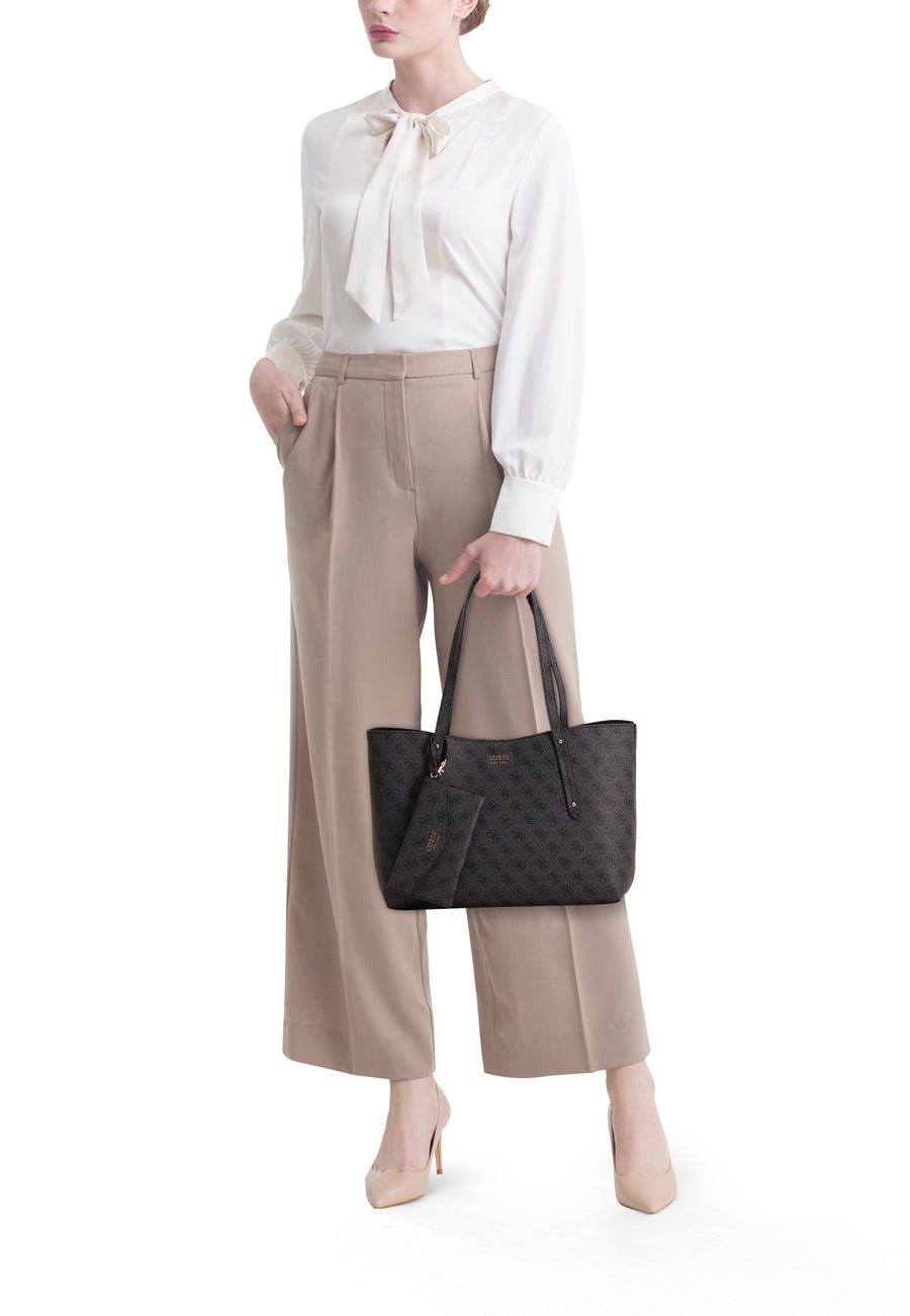Sac à bandoulière noir par Guess, avec motif monogramme, porté avec un chemisier blanc et un pantalon beige.
