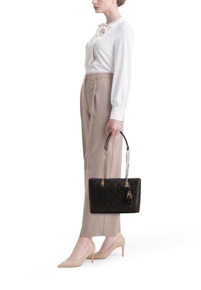 Sac à bandoulière noir de Guess, avec motif gaufré et chaîne dorée, porté avec un chemisier et un pantalon beiges.
