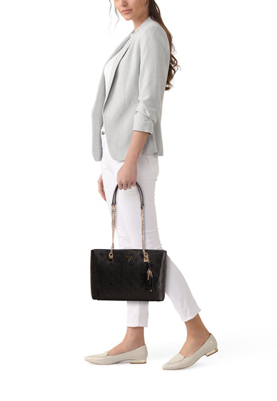 Sac à bandoulière noir Guess avec motif imprimé, porté avec un blazer gris et un pantalon blanc.

