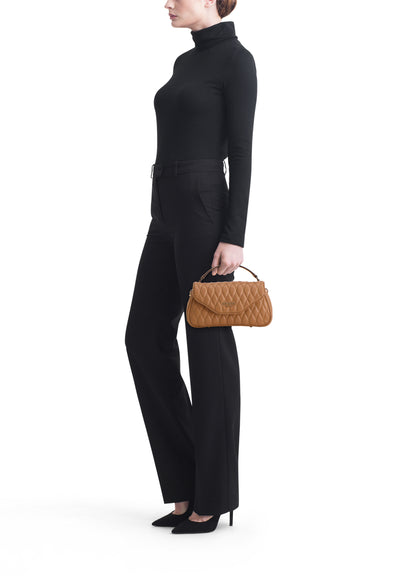 Sac à main beige matelassé de Guess, porté à la main, avec pantalon noir et escarpins pointus.
