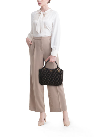 Sac à main noir de Guess avec motif matelassé losange, porté avec un chemisier blanc à lavallière et un pantalon beige.