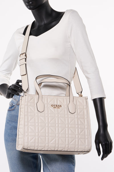 Sac à main écru de Guess, avec motif matelassé et bandoulière ajustable, porté avec un pull blanc et un jean.
