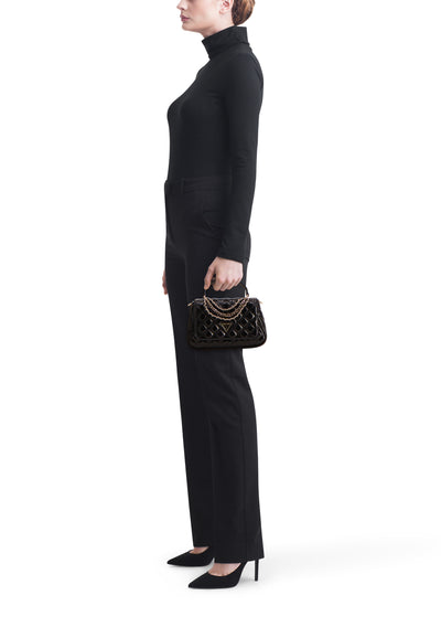 Sac à bandoulière noir de Guess avec chaîne dorée, porté avec un col roulé noir et un pantalon de tailleur noir.