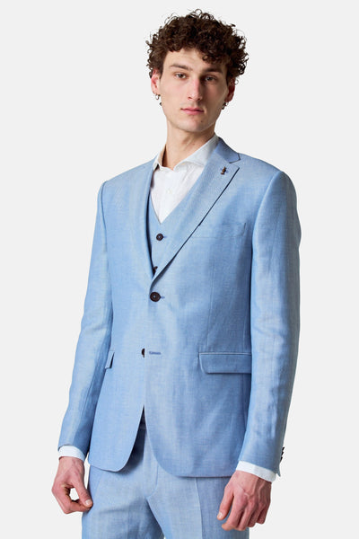 Blazer bleu van Le Fabuleux Marcel de Bruxelles, porté avec un gilet et une chemise blanche, sur fond uni.
