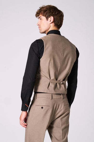 Gilet beige de Le Fabuleux Marcel de Bruxelles, avec patte de serrage au dos et porté sur une chemise noire et un pantalon assorti.