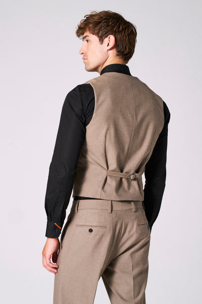 Gilet beige de Le Fabuleux Marcel de Bruxelles, avec patte de serrage au dos et porté sur une chemise noire et un pantalon assorti.