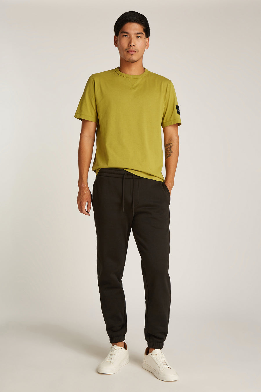 Okerkleurig T-shirt met korte mouwen van Calvin Klein, gedragen met een zwarte joggingbroek en witte sneakers.