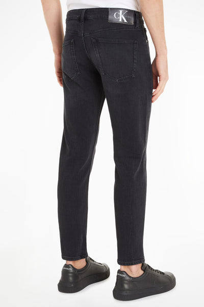 Jean fuselé gris foncé de Calvin Klein, avec une étiquette de logo en cuir à l'arrière et des chaussures noires.