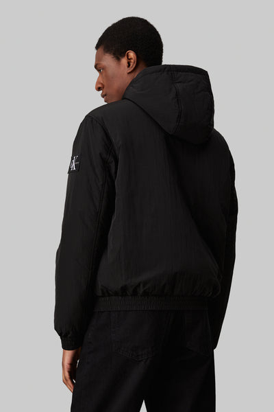 Imperméable noir de Calvin Klein avec capuche et logo sur la manche, porté avec un pantalon noir.
