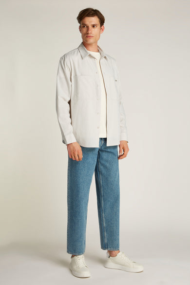 Chemise écrue à manches longues de Calvin Klein, portée ouverte sur un t-shirt, avec un jean bleu et des baskets blanches.