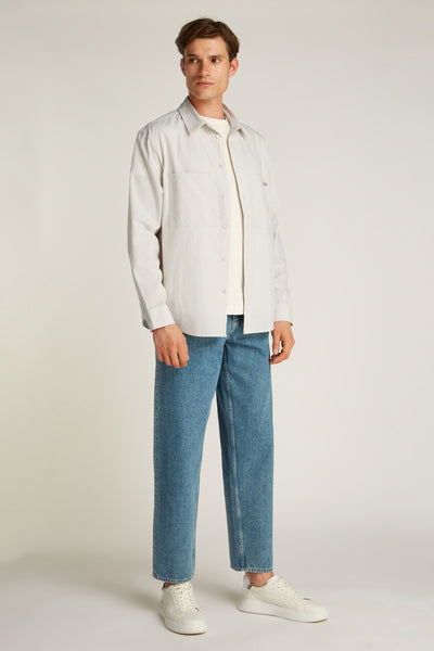 Chemise écrue à manches longues de Calvin Klein, portée ouverte sur un t-shirt, avec un jean bleu et des baskets blanches.