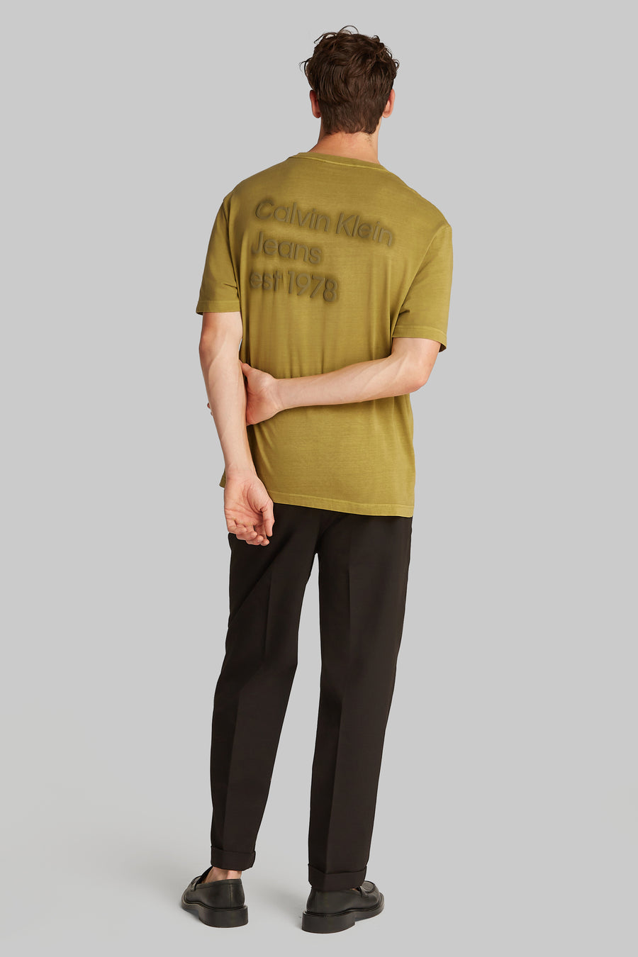 Okerkleurig T-shirt met korte mouwen van Calvin Klein met het merklogo op de rug, gedragen met een zwarte broek.