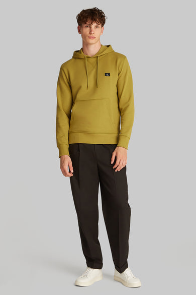 Sweat à capuche ocre de Calvin Klein, avec poche kangourou et logo noir discret, porté avec un pantalon noir et des baskets blanches.
