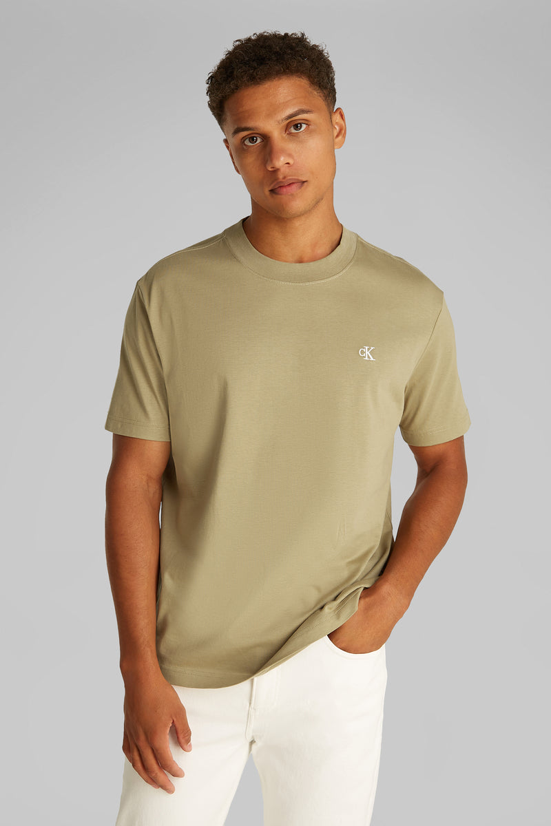 Beige T-shirt met korte mouwen van Calvin Klein, gedragen met een witte broek, hand in de zak.