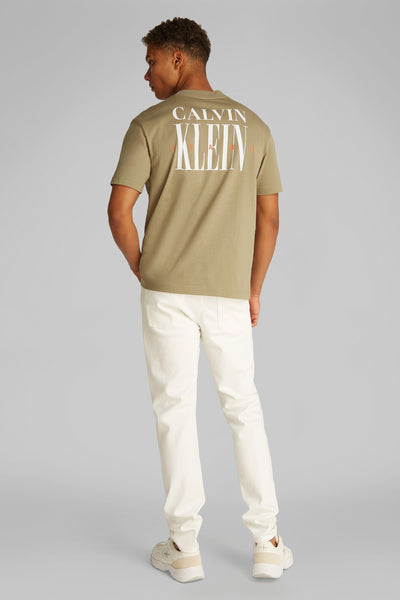 Beige T-shirt met korte mouwen van Calvin Klein, met logo op de rug gedrukt, gedragen met een witte broek.