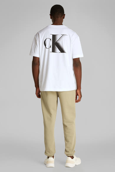 Wit T-shirt met korte mouwen van Calvin Klein, met logo op de rug, gedragen met een beige broek en witte sneakers.