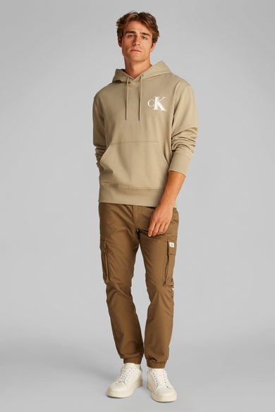 Beige hoodie van Calvin Klein, met logo op de voorkant, gedragen met een bruine cargobroek en witte sneakers.