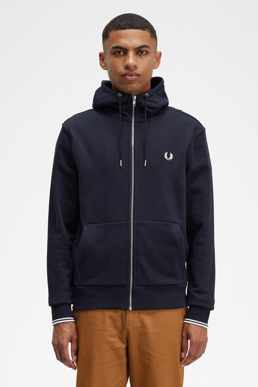 Marineblauwe bodywarmer van FRED PERRY met zilveren ritssluiting en geborduurd logo, gedragen over een bruine broek.