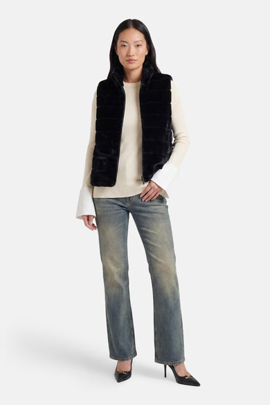 Veste sans manches noire en fausse fourrure d'Access®, portée sur un pull en maille écru à poignets évasés, un jean délavé et des talons noirs.