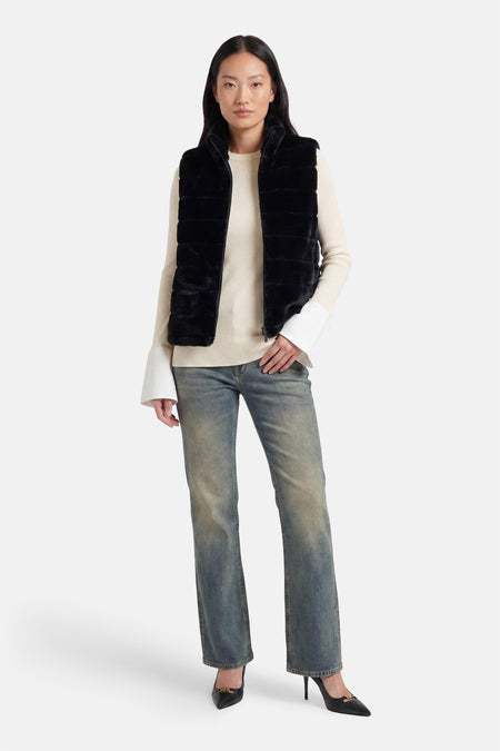 Veste sans manches noire en fausse fourrure d'Access®, portée sur un pull en maille écru à poignets évasés, un jean délavé et des talons noirs.