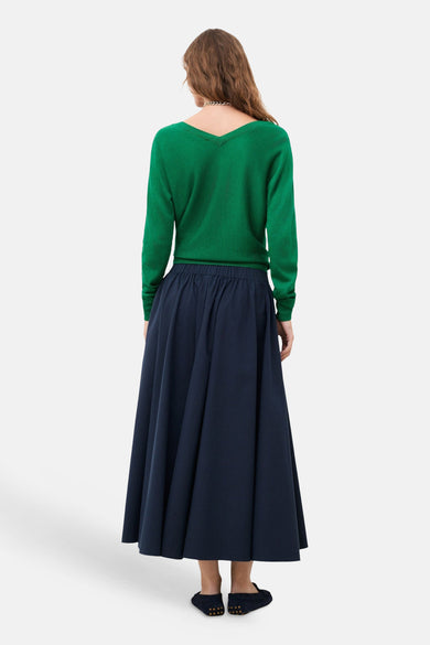 Jupe longue bleue de CAROLL, tombant jusqu'aux chevilles, portée avec un pull vert et des chaussures plates.
