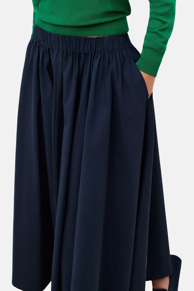 Jupe longue bleue par CAROLL avec taille élastique, ample et drapée, portée avec un pull vert.
