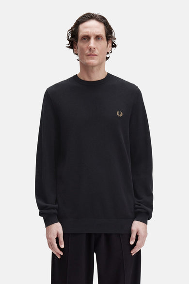 Zwarte rondehals trui van Fred Perry, versierd met een geborduurd logo op de borst, gedragen met een zwarte broek.