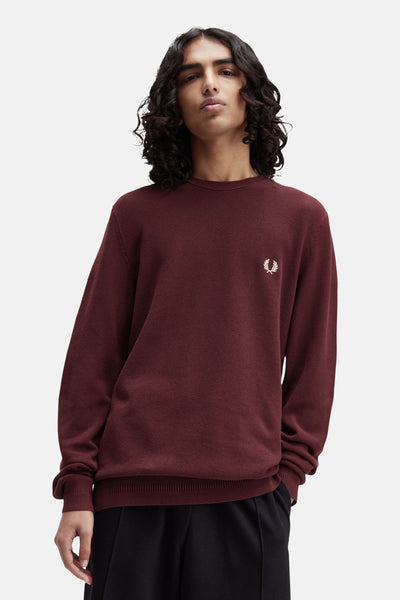 Pull à col rond bordeaux de FRED PERRY, avec logo brodé, porté sur un pantalon noir.

