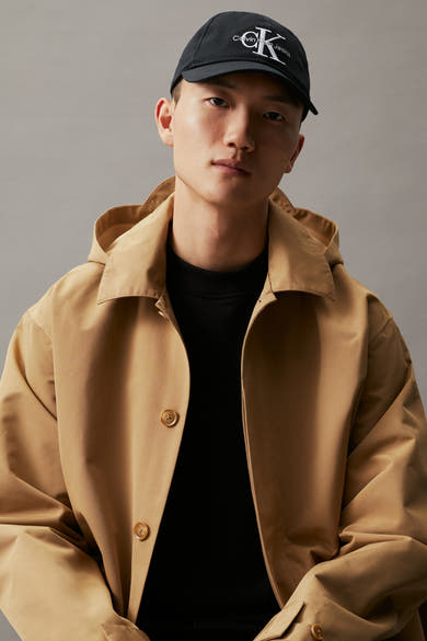 Casquette noire de Calvin Klein Jeans, avec un logo brodé sur le devant et un homme portant un trench-coat beige.
