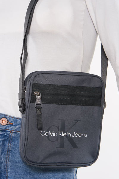 Sac à bandoulière gris de Calvin Klein Jeans, porté sur le corps avec un t-shirt blanc et un jean.
