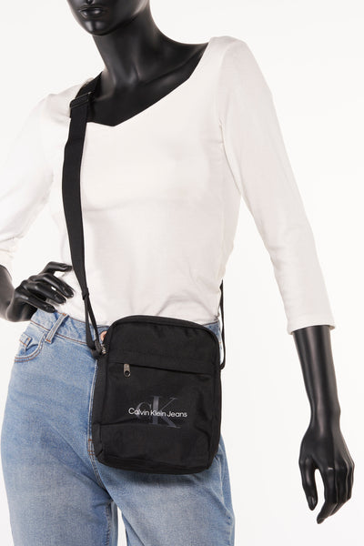 Zwarte schoudertas van Calvin Klein Jeans gedragen door een model met wit overhemd en blauwe jeans.