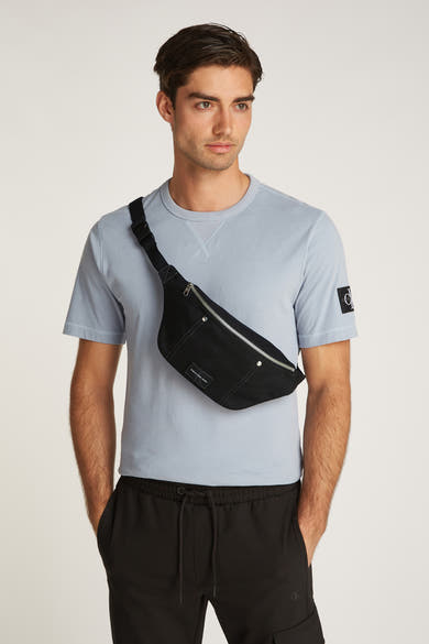Zwarte Calvin Klein schoudertas, gedragen over het lichaam, met een lichtblauw overhemd en een zwarte broek.