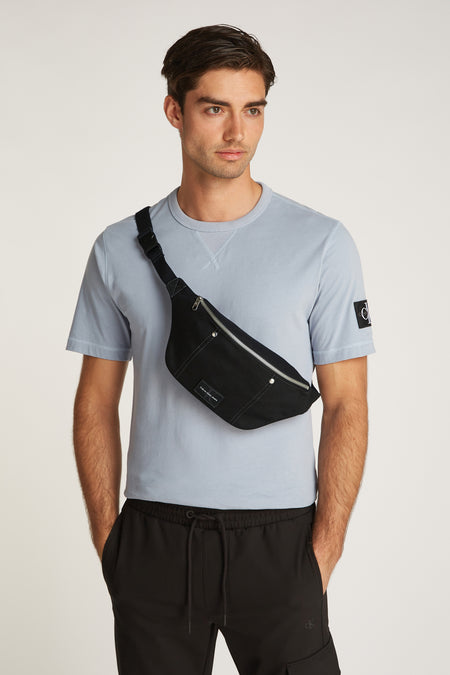 Sac à bandoulière noir de Calvin Klein, porté sur le corps, avec une chemise bleu clair et un pantalon noir.