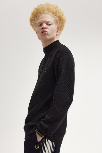 Pull à col montant noir de Fred Perry, texture gaufrée visible, porté avec un pantalon à rayures.