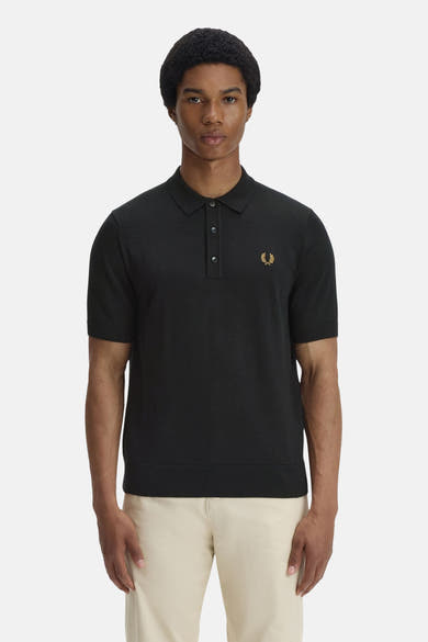 Polo noir de FRED PERRY, orné d'un logo brodé doré sur la poitrine, porté avec un pantalon beige.
