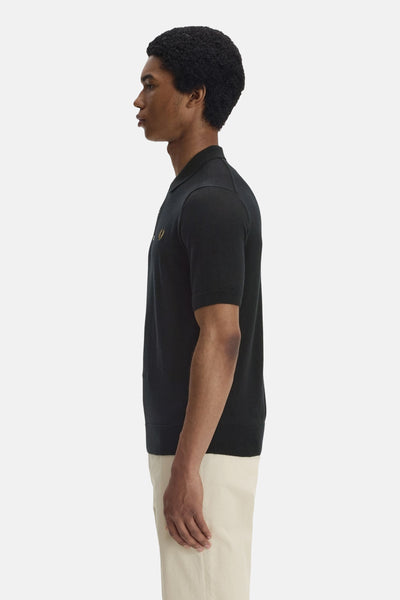 Polo noir par Fred Perry, avec logo brodé couleur or, porté avec un pantalon beige, vue de côté.
