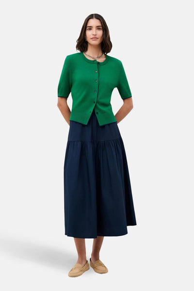 Cardigan vert en maille by CAROLL, porté avec une jupe longue bleu marine et des chaussures plates.